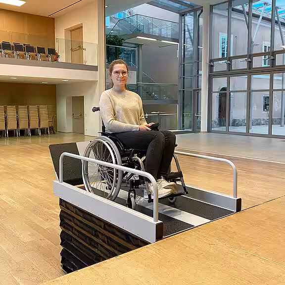 Eine Frau im Rollstuhl fährt auf eine Plattformliftrampe im Innenraum zu, nahe einer Treppe. Im Hintergrund stehen Spinde und ein Laufband.