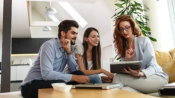 Drei Personen sitzen auf einem Sofa in einem modernen Wohnzimmer und schauen gemeinsam auf ein Tablet, während sie sich beraten. 
