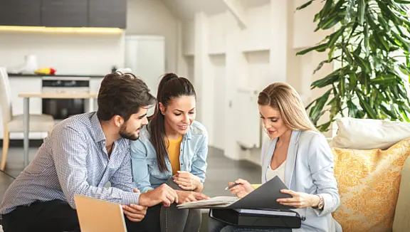 Drei Menschen sitzen an einem Tisch im Wohnzimmer, schauen gemeinsam in einen Ordner und besprechen Unterlagen. Im Hintergrund ist eine Küche sichtbar.