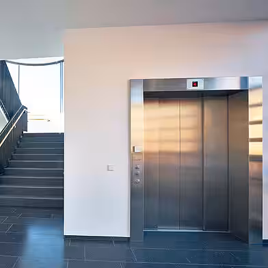 Ein älterer Mann und eine ältere Frau sitzen an einem Tisch und geben einem jüngeren Mann freundlich die Hand; im Hintergrund sieht man ein Regal mit Büchern.