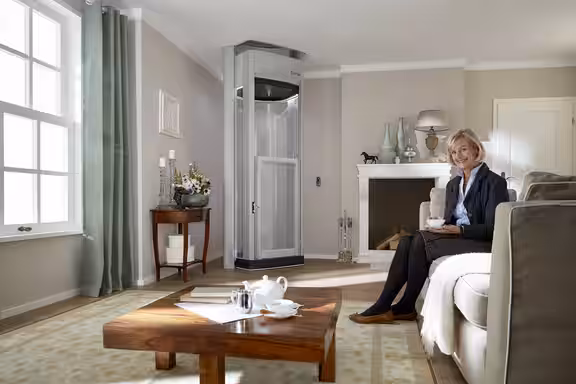 Eine ältere Dame sitzt lächelnd auf der Couch. Im Hintergrund ist ein LIFTON DUO Homelift.