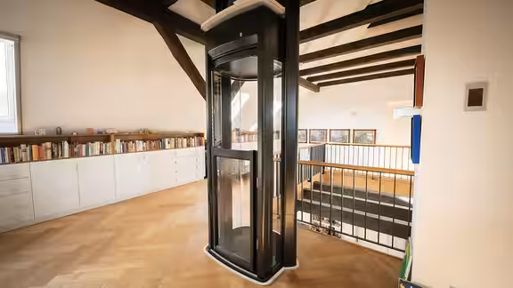 Ein moderner Homelift steht in einem großzügigen, hellen Wohnraum mit Bücherregal, Holzfußboden und sichtbaren Holzbalken, umgeben von einer Galerie mit Geländer.