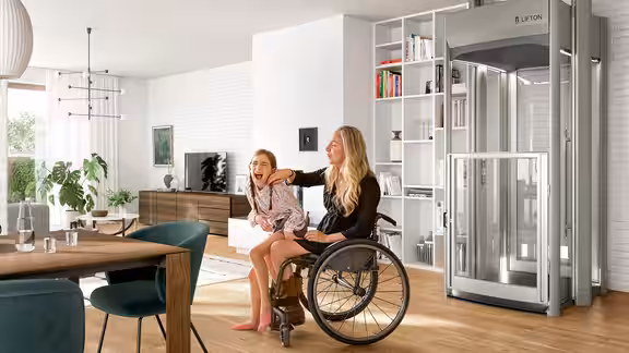 Zwei Personen sitzen lachend im Wohnzimmer, eine im Rollstuhl, neben ihnen steht ein moderner Homelift von Lifton in einer offenen, hellen Wohnumgebung.