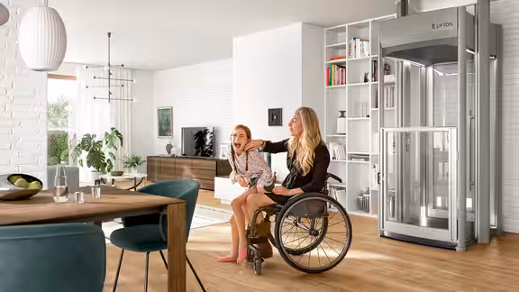 Zwei Personen sitzen lachend im Wohnzimmer, eine im Rollstuhl, neben ihnen steht ein moderner Homelift von Lifton in einer offenen, hellen Wohnumgebung.