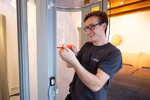 Ein Techniker montiert lächelnd mit einem Schraubendreher Teile an einem Homelift in einem hellen, wohnlichen Raum mit Holzbalkendecke und moderner Einrichtung.