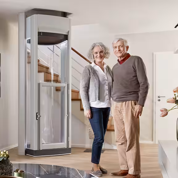 Ein älteres Paar steht lächelnd in einem modernen Wohnzimmer neben einem Homelift; im Hintergrund befindet sich eine Treppe und die Atmosphäre wirkt freundlich und hell.