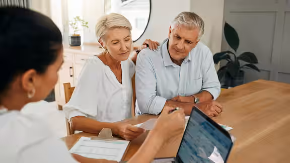 Ein älteres Paar sitzt an einem Holztisch, betrachtet gemeinsam mit einer Homelift-Beraterin Unterlagen und einen Laptop, freundlich und aufmerksam im modernen, hellen Raum.