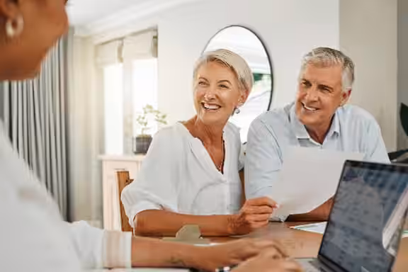 Ein älteres Paar sitzt lächelnd am Tisch, hält ein Papier und spricht mit einer Person, die vor einem Laptop sitzt; helle, freundliche Wohnumgebung.
