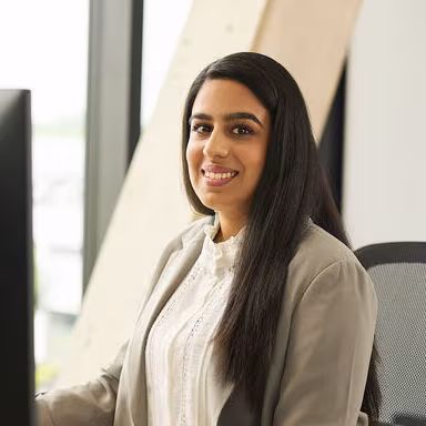 Eine Homelift-Beraterin sitzt an einem Schreibtisch, schaut lächelnd in Richtung Kamera, trägt eine weiße Bluse und einen grauen Blazer, im Hintergrund ist ein Fenster mit Tageslicht.