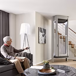 Ein älterer Mann sitzt auf der Couch und nutzt die LIFTON Homelift Fernbedienung. Im Hintergrund fährt der LIFTON Homelift runter